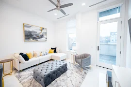 Furnished living room with white walls and floors, glass patio door to balcony, tall window with shades, and ceiling fan