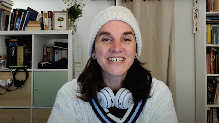 Still frame of Tea Uglow in a beanie with headphones around her neck with a curtain and bookcase in the background.