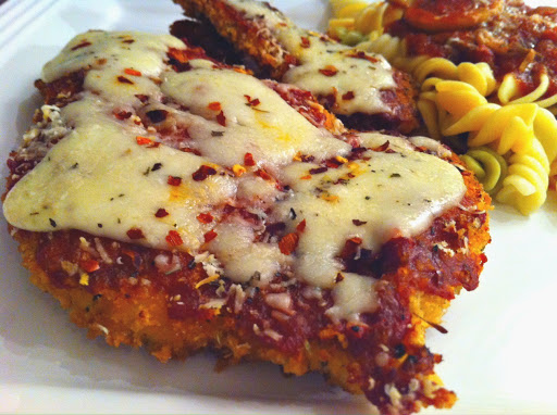 A chicken parmesan topped with melted cheese and red pepper flakes sitting on a white plate with pasta in the background.