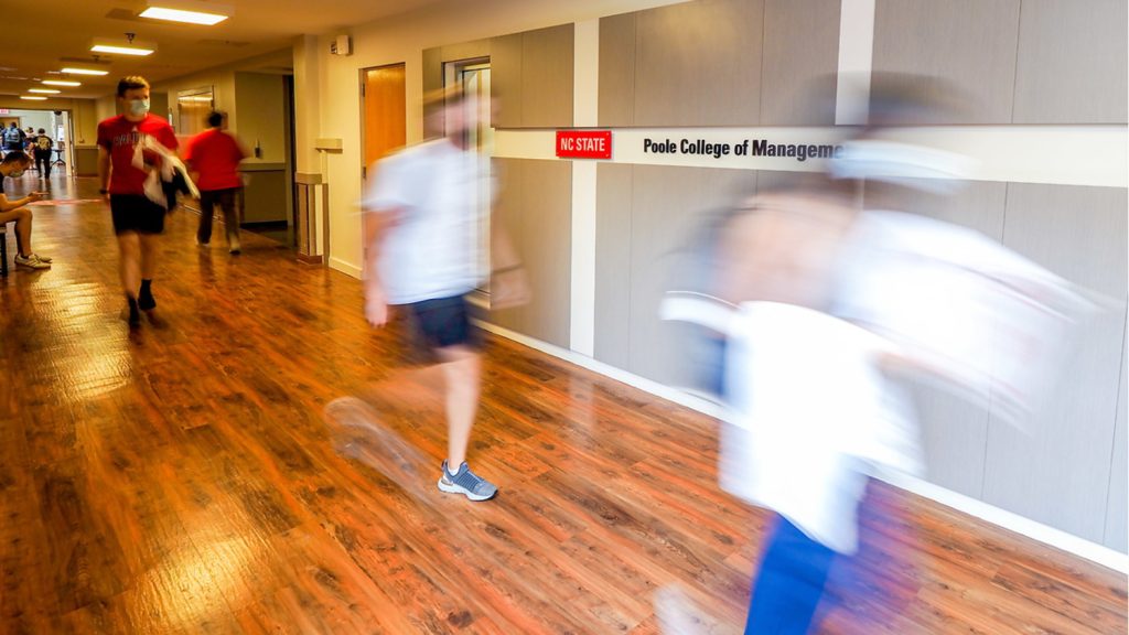 Out-of-focus students walk past a wall with a "NC State Poole College of Management" sign