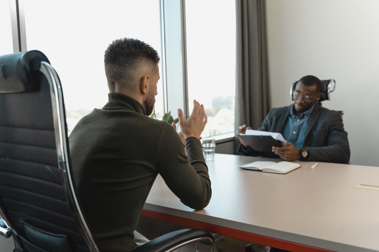 A imagem mostra dois homens negros em uma sala de reunião. Eles estão de frente para o outro e são separados por uma mesa com tampo cinza. No lado esquerdo da foto, está um homem jovem, ele possui cabelo crespo no topo da cabeça e as laterais estão raspadas. Em frente a ele, está um homem negro que aparenta ser um pouco mais velho. Ele veste terno, usa óculos e faz anotações em um papel que está apoiado em uma prancheta, que está na mão esquerda dele. 