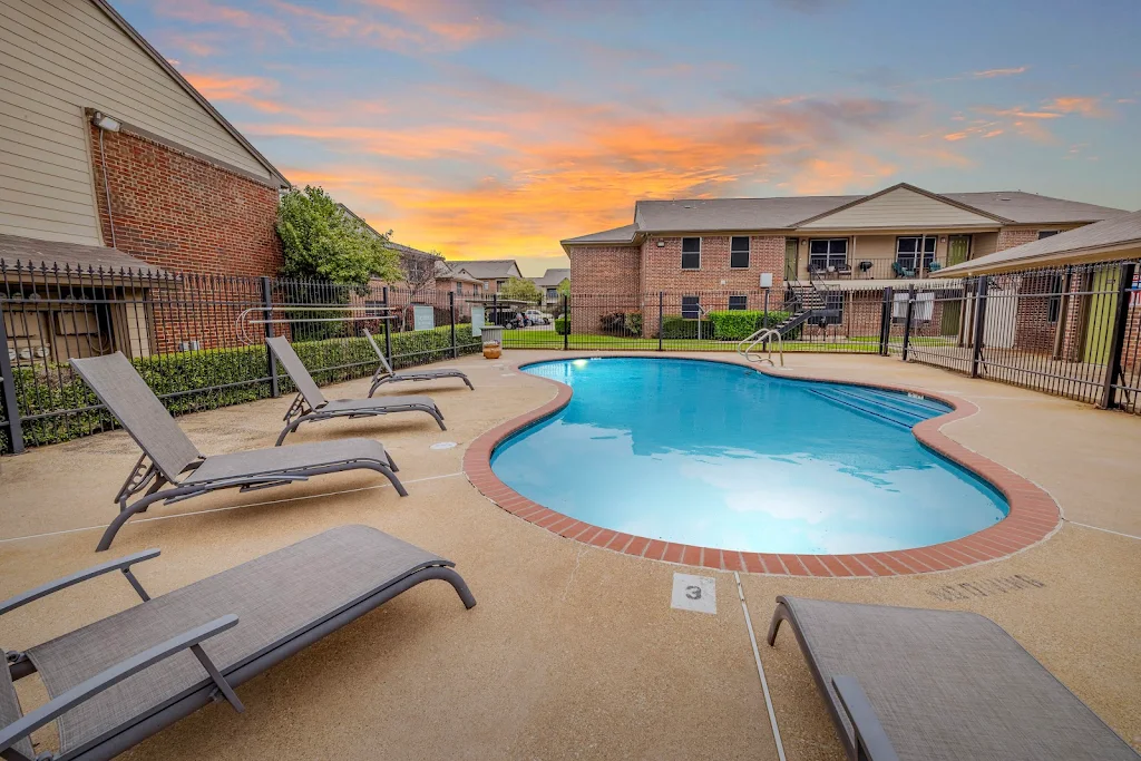 Apartment complex pool surrounded by lounge chairs at sunset with brick buildings in the background.