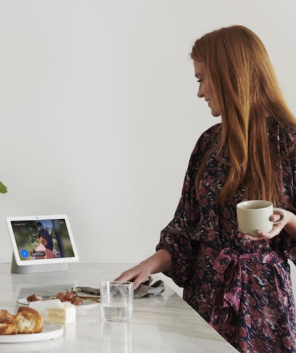 A woman drinking coffee looks at a Nest Hub display on a table.