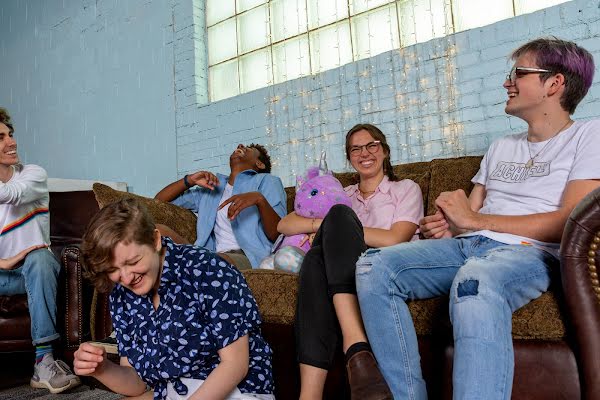 Un grupo de jóvenes riendo y sonriendo sentados juntos en un sofá. Uno de ellos sostiene un unicornio morado de peluche. Hay luces centelleantes colgadas contra la pared de ladrillo pintado.