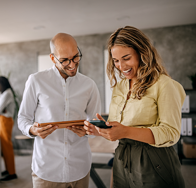 A male wearing a white shirt reviewing a work project on mobile tablets with a female colleague in a yellow shirt while standing in their office.