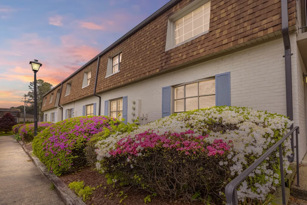 Twilight view of apartment complex with gabled roofs, flowering shrubs, and a lit pathway lamp.