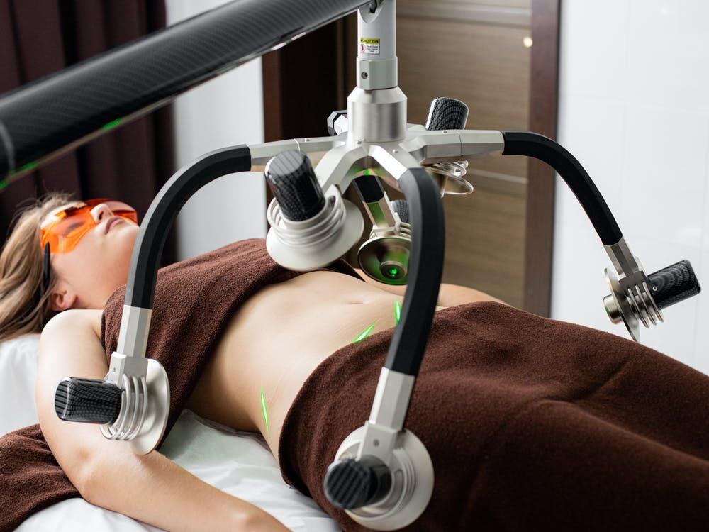 Free Female client lying on table under lasers of modern weight loss machine on belly during liposuction procedure in medical clinic Stock Photo