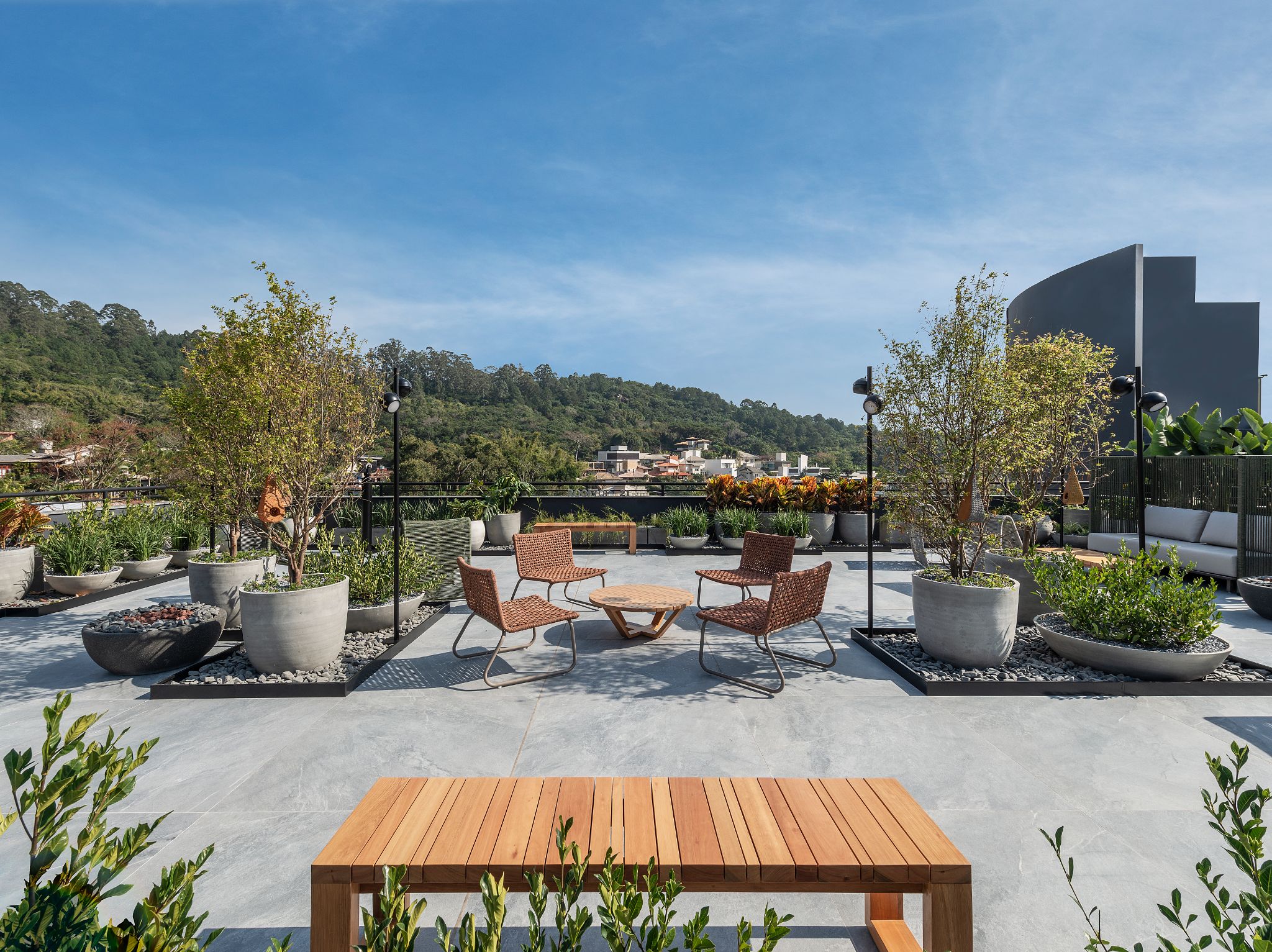 Casacor Santa Catarina - jardim a céu aberto com piso em cimento queimado, bancos de madeira, diversos vasos de plantas, mesinha redonda de madeira com quatro cadeiras amadeiradas.