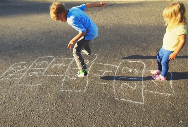 two children playing hopscotch outside on a sidewalk. The hopscotch is written in chalk from 1-10.