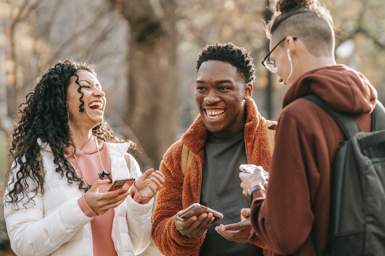 A imagem mostra três amigos reunidos e sorrindo, como em uma conversa informal. Da esquerda para a direita, é possível ver uma jovem branca, de cabelos cacheados pretos na altura do peito e que está segurando um celular com a mão direita. Ao lado dela, há um rapaz negro, de cabelos crespos curtos e que também segura um celular com a mão esquerda. Por fim, há um rapaz branco, com as laterais da cabeça raspadas e a parte do meio do cabelo é comprida, mas os cabelos estão presos em um coque. Ele está de costas para a imagem e é possível vê-lo mexendo no celular. 