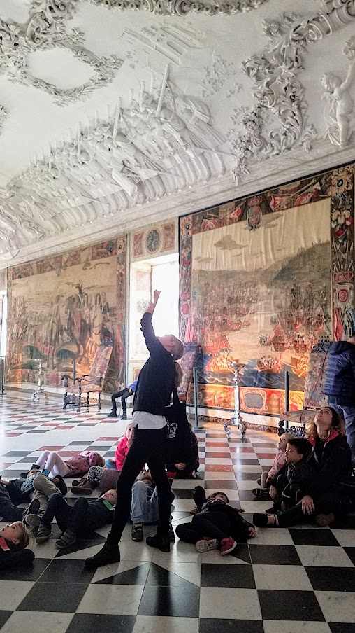 Visiting Rosenborg Castle in Copenhagen. On the third floor and is where you'll find the impressive Throne Room, also known as the Long Hall or Knights’ Hall. It was intended as a ballroom, and here you will find the coronation thrones for the king and queen and three life-size silver lions standing guard. On the other side of the room is displayed the throne for audience. In this room I caught back up with the kids group who were lying down getting a lesson on the ceiling.