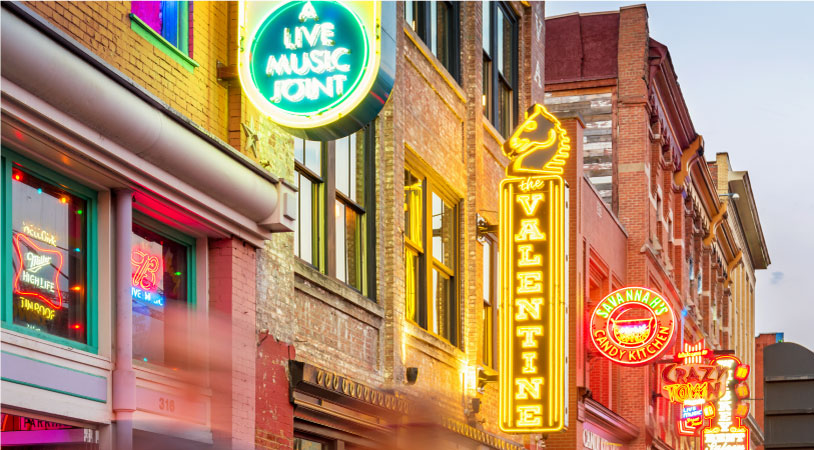 Dusk sets in over downtown Nashville, Tennessee. Brightly lit signs hang from tall brick buildings inviting visitors to come inside for live music.