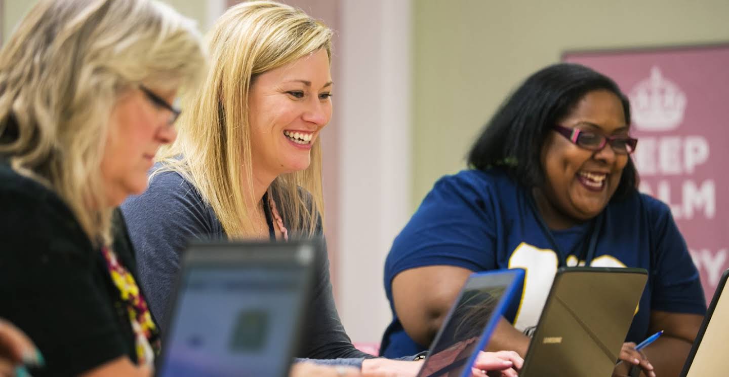 Três educadoras estão sorrindo sentadas olhando para seus laptops.