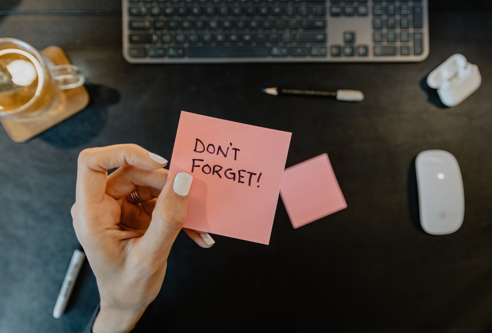 On a dark wood table lays a cup of coffee, and the keyboard to a laptop. In front of it, is a pair of hands holding a bright pink sticky note titled "Don't Forget!"