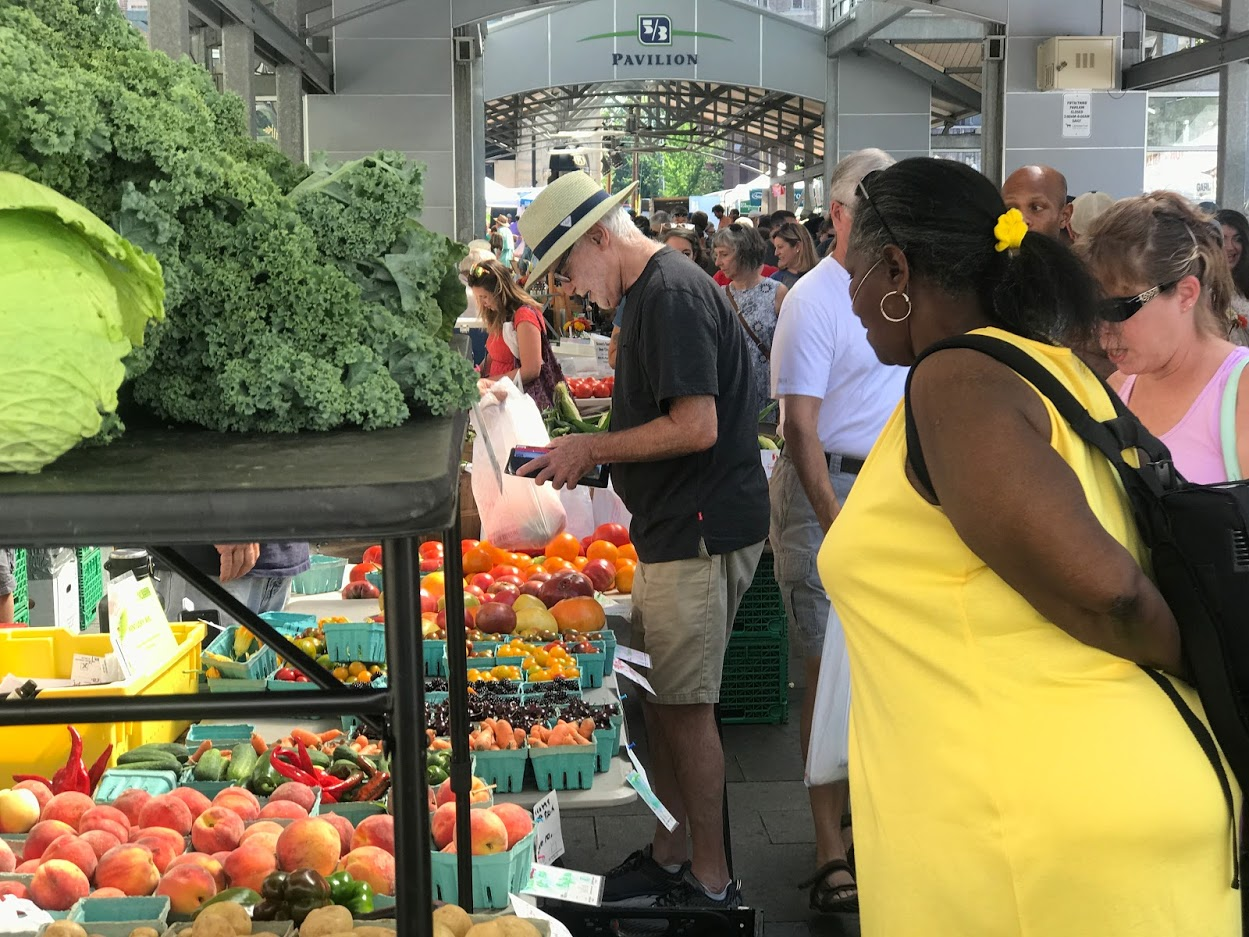 Woman in yellow looks at seasonal produce in a crowded farmers market.