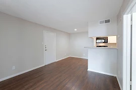 Living and dining rooms with dark wood floors, unit entry door, cream walls, white trim, bar top into kitchen