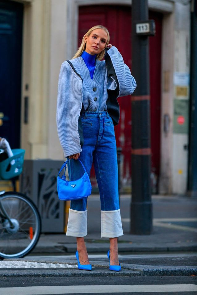 Mulher branca e loira, com um look monocromático azul. Ela usa calça jeans, blusa de lã e uma blusa de gola alta. Está em um cenário urbano.