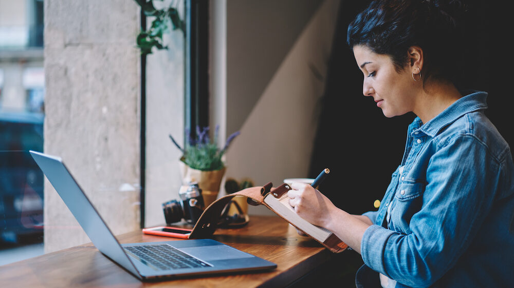 Caucasian female blogger writing content information for own website while sitting with laptop device at cafeteria table, attractive woman preparing to course work indoors holding textbook