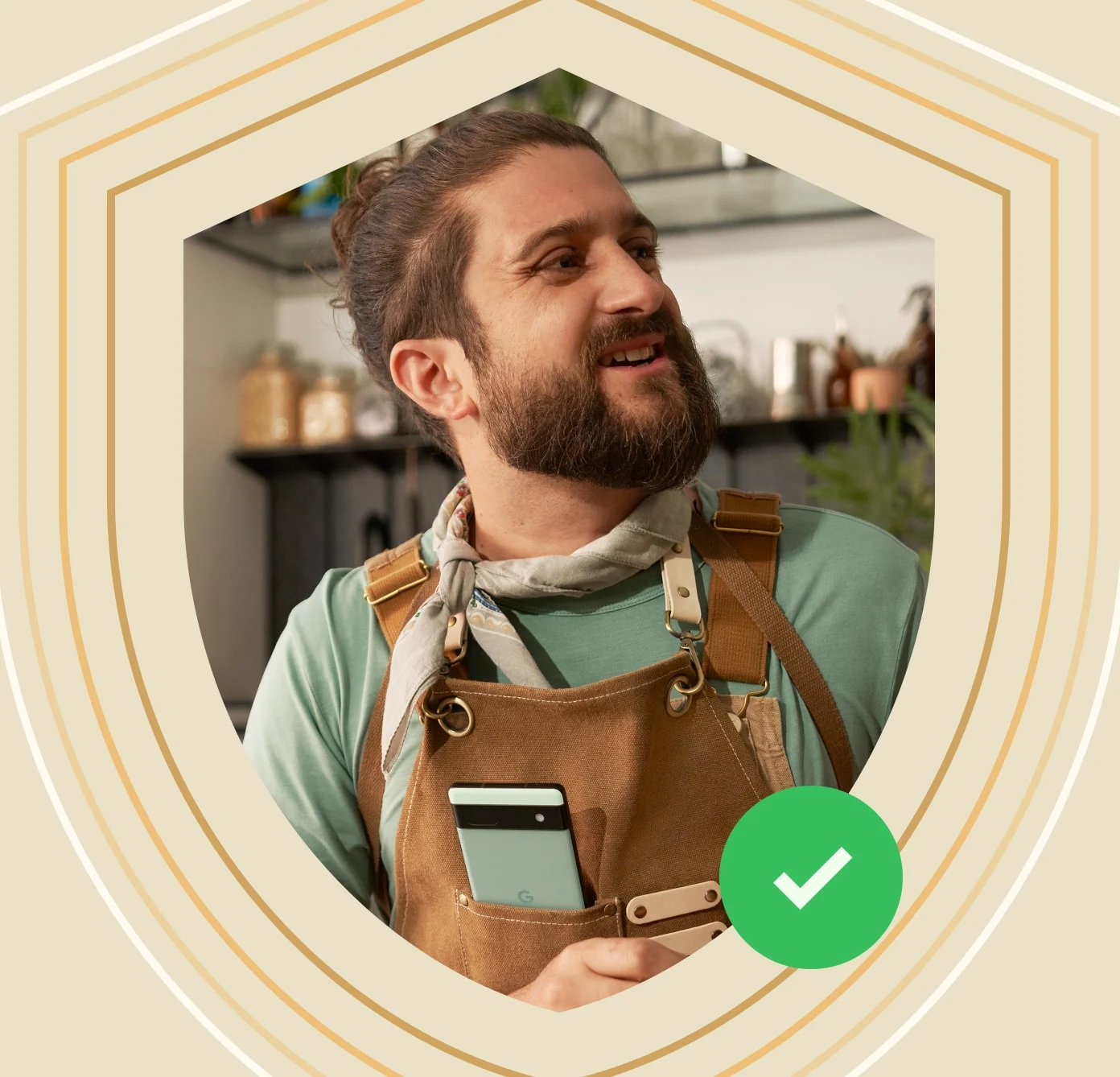 An artist smiles with a Sage green Pixel 6a in the pocket of their smock, with a border of a security shield and a green checkmark