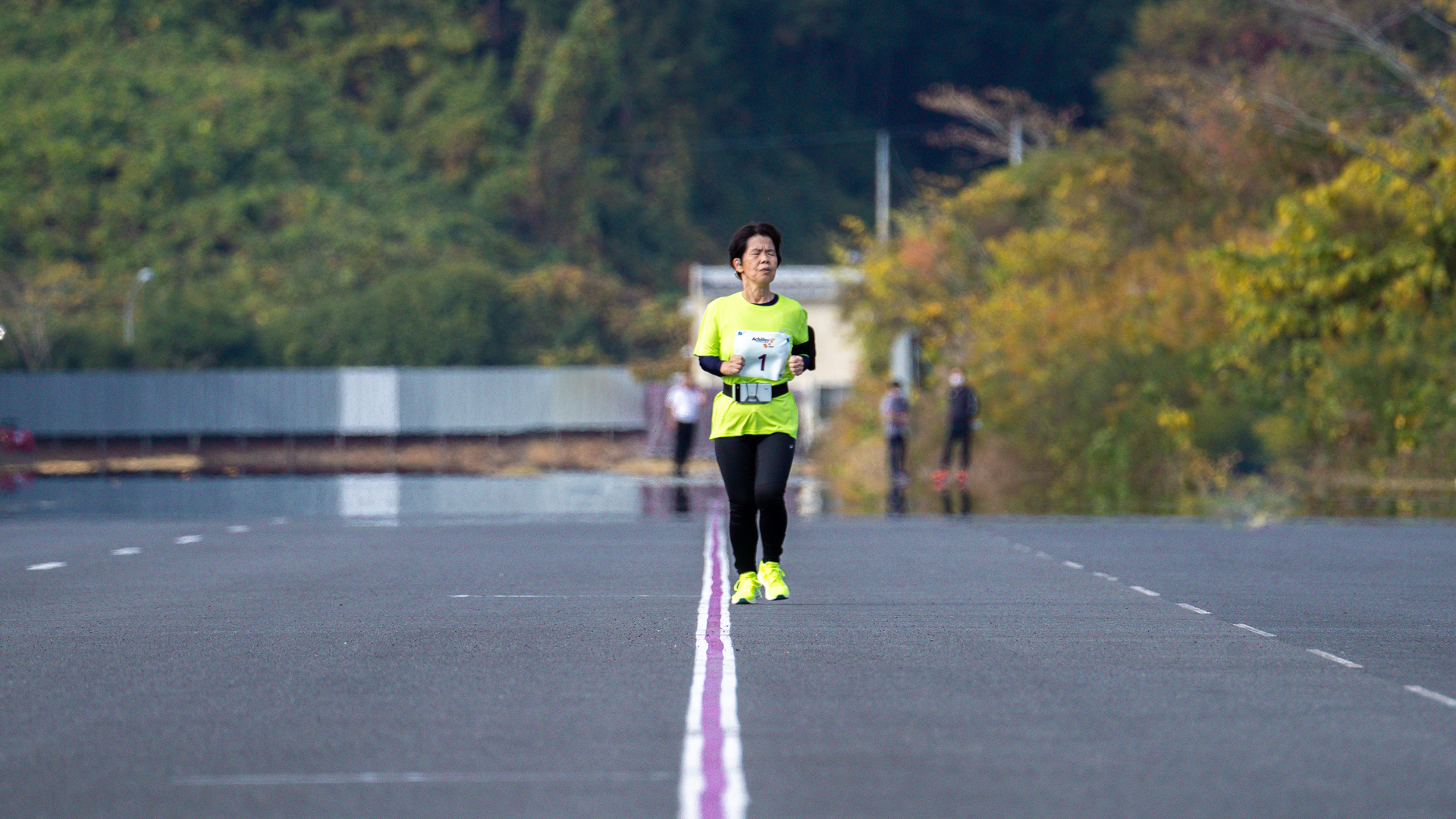 Purple tape is stretched in a straight line across a large area surrounded by trees. A woman with a smartphone strapped to her waist and an Ekiden race bib with the number "1" on it is running along the purple line..