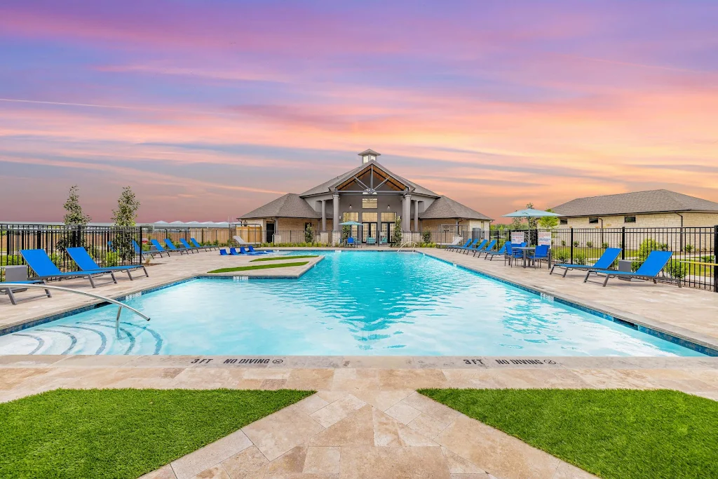 Community swimming pool surrounded by blue lounge chairs and a gated fence with the clubhouse in the background at dusk