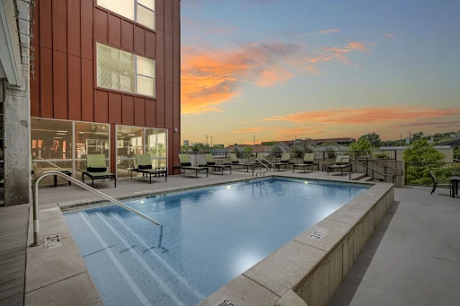 Apartment complex with a refreshing pool and lounge area, surrounded by scenic views and a vibrant sunset sky.