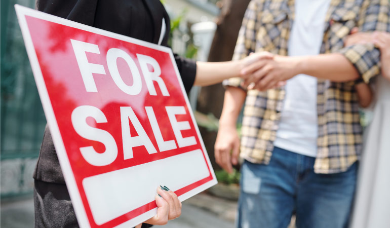 A realtor holding a “for sale” sign and congratulating a couple on the purchase of their new home