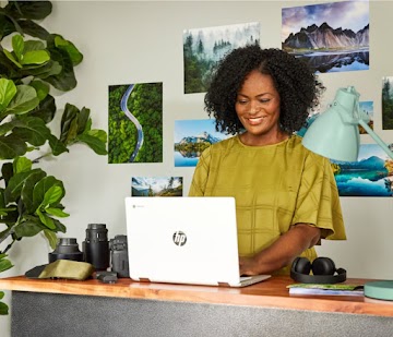 Una persona parada frente a una pared con fotos escribe en un Chromebook que está apoyado en una mesa rodeada de objetivos para cámaras.