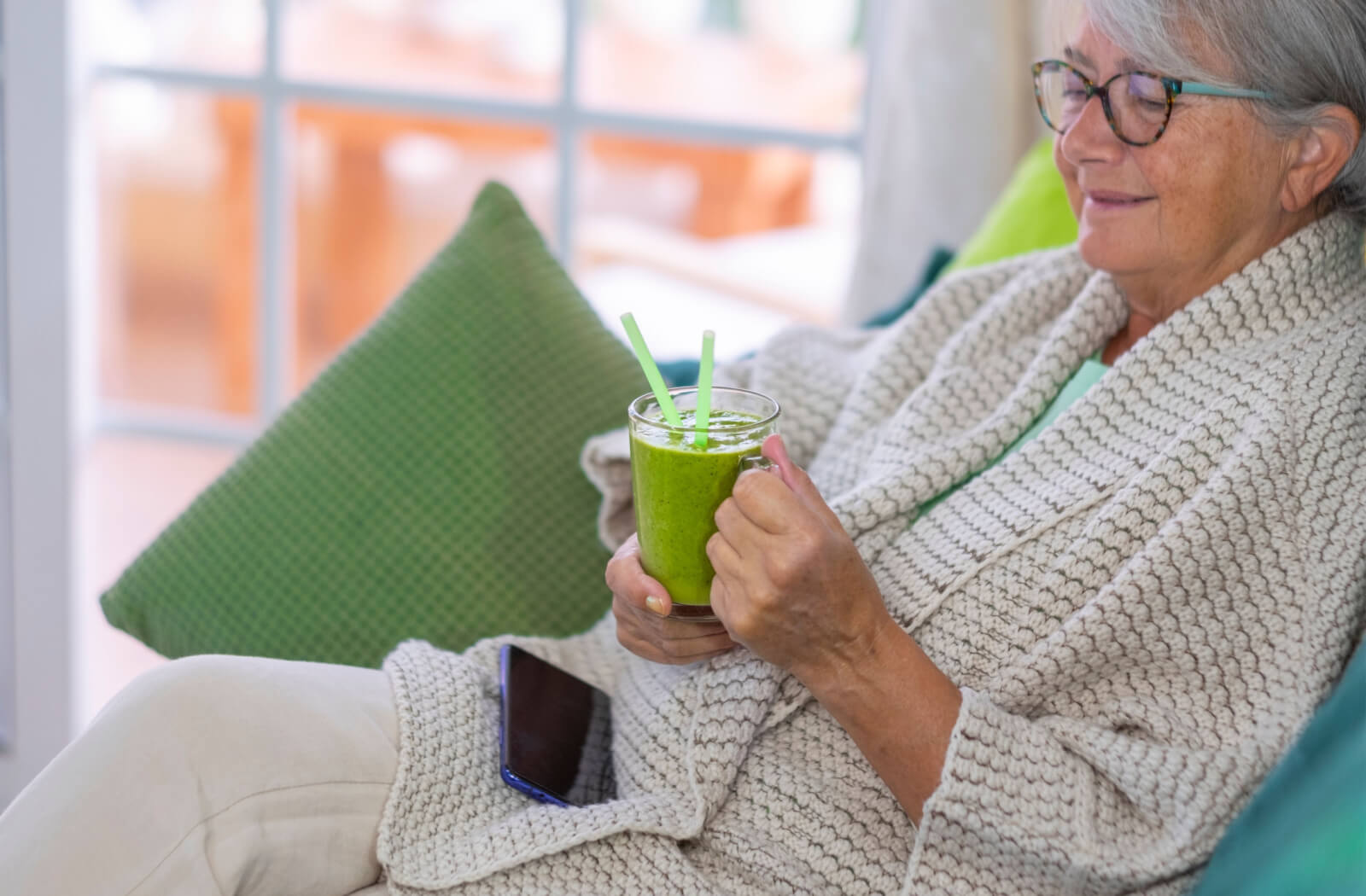 A senior woman in a knitted sweater relaxing and enjoying a smoothie.