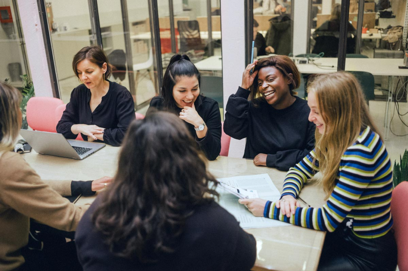 A group of women sit at a table working on their laptops, smiling and laughing.