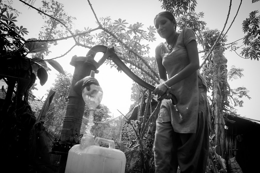 Description of . “In the camp, we had to follow a schedule to get water in the morning and evening. This woman is getting water for cooking and drinking. We stand in line to get water. Sometimes we wait one hour. America is very different from Nepal. This country is very fast. Very easy.  When I arrived at the Los Angeles airport I went to restroom. I put my hands under the faucet and the water came. How is it? I felt like someone was giving from above. Chaka ban! Surprised! The air dryer also surprised me. At that time a stranger taught me to dry my hands. I used the hand dryer and it was loud. I was afraid.”    - Mohan Rai