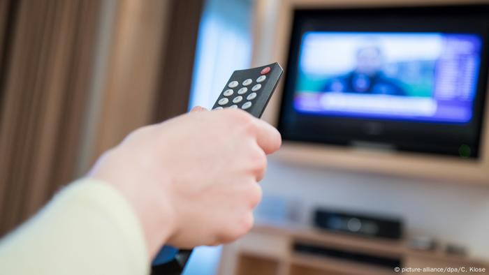 Mão de telespectador diante de TV, com controle remoto