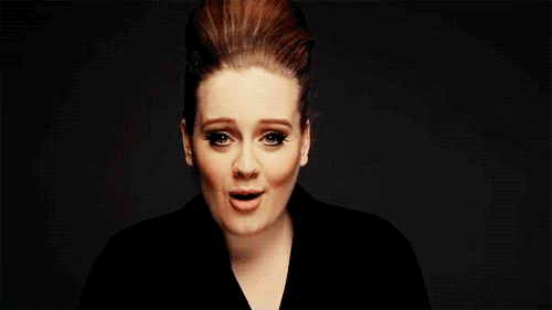 [Image is an animated gif of Adele from the shoulders up. What can be seen of her clothing is black, almost blending into the background, and with a mild v-neck down it. She is looking straight ahead, saying something, before she turns her whole body and face to the left side and smiles.]
