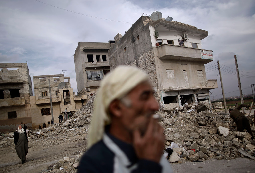 Description of . In this Wednesday, Dec. 12, 2012 file photo, an elderly Syrian man smokes a cigarette as he stands next to a residential building destroyed in a government airstrike, in Maaret Misreen, near Idlib, Syria. A U.S.-based rights group on Thursday accused Syria of war crimes by indiscriminate and sometimes deliberate airstrikes against civilians, killing at least 4,300 people since last summer. (AP Photo/Muhammed Muheisen)