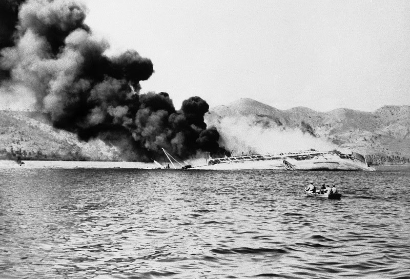 Description of  August 3, 1942: After hammering Port Moresby for two days, Japanese bombers finally sank this Australian transport which sends up a cloud of smoke. She drifted onto a reef and heeled over. Flaming oil can be seen at left. The men in a small boat, foreground, are looking for victims. (AP Photo)