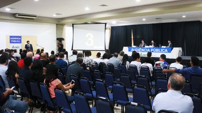 Foto da audiÃªncia pÃºblica mostrando a mesa principal e o pÃºblico presente. 
