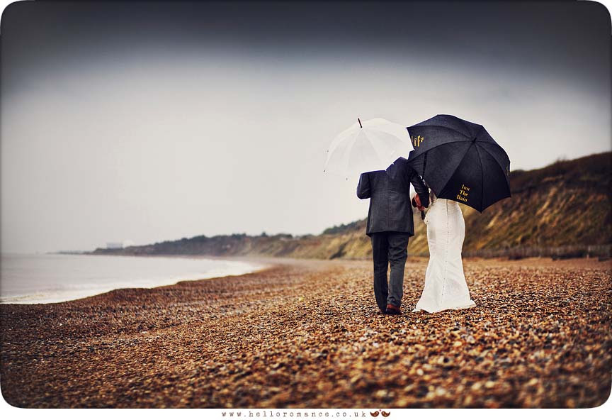 Bride and Groom walking in rain on Dunwich Beach with umbrellas - Westleton Crown Wedding Photography - Kate and Rob - Hello Romance Wedding Photography Suffolk