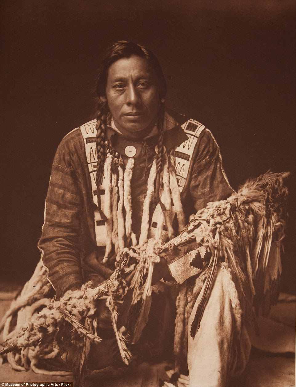 A Piegan man kneels holding medicine pipe described by Curtis as 'long pipe-stems variously decorated with beads, paint, feathers, and fur'