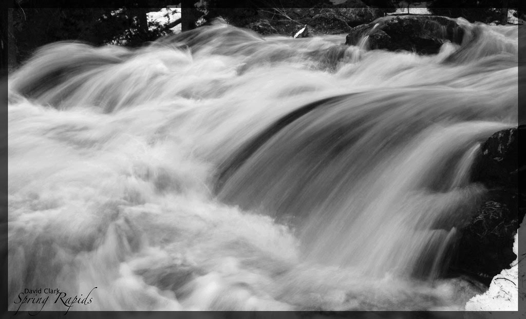 A black and white photo of a roaring river going over a small waterfall, with much foam and streaks of water.