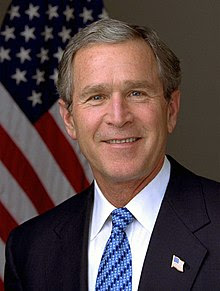 A portrait shot of a smiling older male looking straight ahead. He has short gray hair, and is wearing a dark navy blazer with a blue styled tie over a white collared shirt. In the background is an American flag hanging from a flagpole.