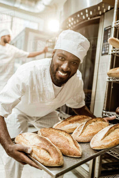 Happy african american baker taking bread loaves from oven at baking