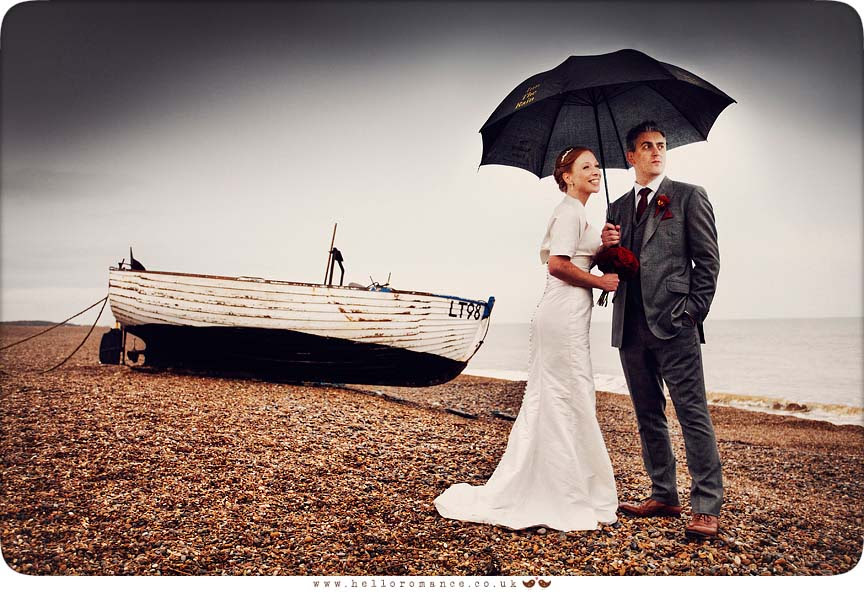 Bride and Groom in rain on Dunwich Beach with umbrellas with boat dark sky- Westleton Crown Wedding Photography - Kate and Rob - Hello Romance Wedding Photography Suffolk
