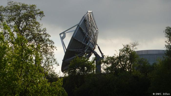 An antenna flanked by trees and bushes against a dark gray backdrop of clouds.