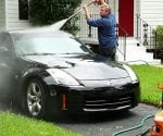 Man using a pressure washer to clean a black sports car in driveway.