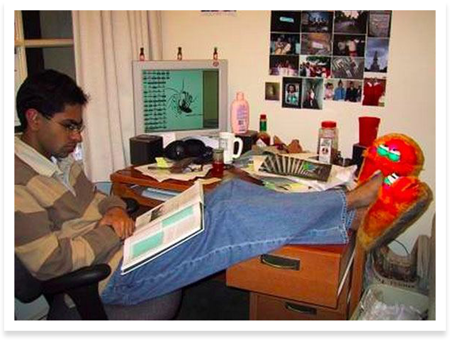 Young, skinny Ramit Sethi sitting in his messy dorm room reading a book. His feet are propped up on his desk and he is wearing fuzzy red slippers.
