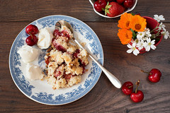 Cherry crumble dessert with strawberries and vanilla cream, served on a blue plate. Cherries, strawberries, flowers behind. Dark wooden background, top view