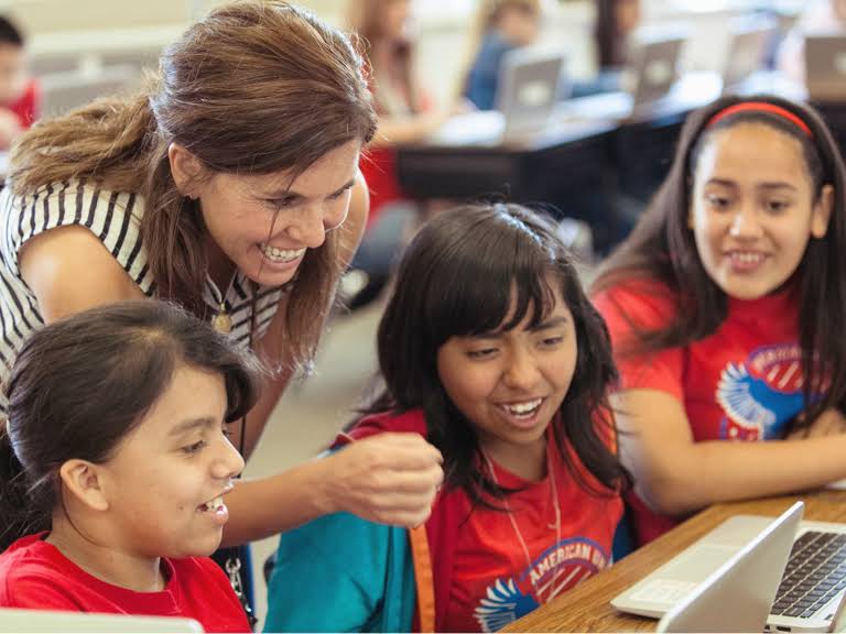 Una profesora sonriente mira algo en una pantalla con tres alumnas que están sentadas.
