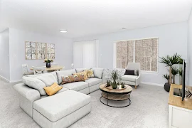 Modern apartment living room with a gray sectional sofa, round coffee table, light blue walls, and decorative plants.