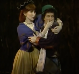 A medium shot of a woman and a man in a green hat. The woman is grabbing the man's face by the chin, as if she has just turned it to face something.