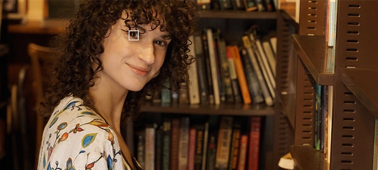 A woman in a library looking towards the camera - a small white square over one of her eyes represents Eye AF for video